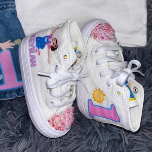 Puede incluir: Zapatillas altas blancas con ilustraciones y adornos personalizados. Un zapato presenta una niña de dibujos animados y el nombre "TEYLAH". El otro tiene un sol y el número "1". Las punteras están adornadas con gemas rosas e iridiscentes.