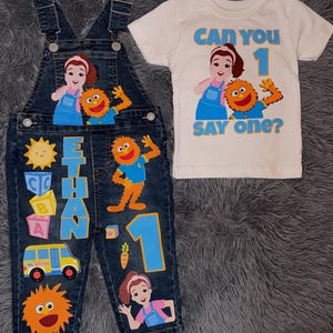 Op de afbeelding: Een spijkerbroek en witte t-shirt set voor een kind. De spijkerbroek heeft kleurrijke cartoon karakter applicaties, de naam "ETHAN" en het nummer "1". Het t-shirt zegt "CAN YOU SAY ONE?" met bijpassende cartoon karakters.