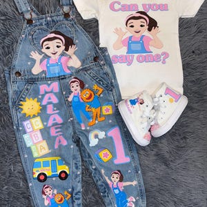 May include: A denim overall set with a matching white t-shirt and sneakers. The overalls feature colorful cartoon graphics, letters, and a school bus. The t-shirt has a cartoon graphic and the text "Can you say one?" The sneakers have a pink number 1.