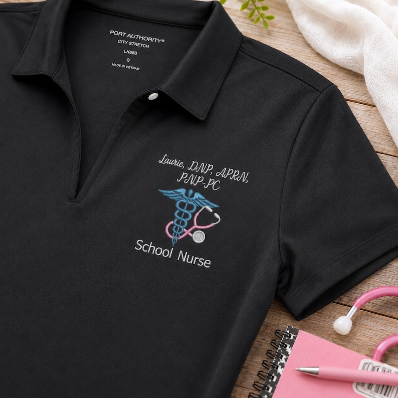 K&ouml;nnte beinhalten: Ein schwarzes Poloshirt mit V-Ausschnitt und kurzen &Auml;rmeln. Auf der Brust befindet sich eine wei&szlig;e Stickerei mit dem Text Laurie, DNP, APRN, PNP-PC und School Nurse sowie ein blaues Caduceus-Symbol mit einem rosa Stethoskop.
