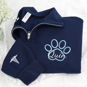 May include: Navy blue quarter-zip sweatshirt with a light blue paw print and the name "Quin" embroidered on the front. The left sleeve cuff features a white embroidered medical symbol. The sweatshirt has a silver zipper.