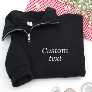 Puede incluir: Sudadera negra con cuello con cremallera y el texto bordado "Custom text" en blanco. La sudadera está doblada, mostrando la cremallera y la tela suave. El fondo incluye un patrón de cuadros rojos y blancos.