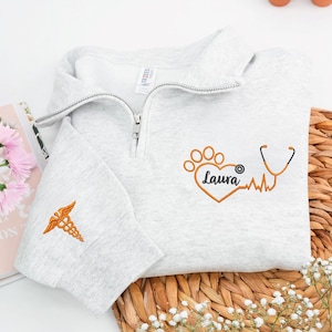 May include: A light gray quarter-zip sweatshirt with an embroidered orange medical caduceus on the left sleeve. The front features an orange paw print heart design with the name "Laura", a stethoscope, and a heartbeat line.