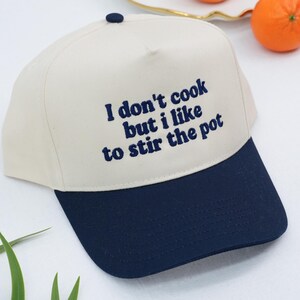 Op de afbeelding: Crèmekleurige baseballcap met een marineblauwe klep. De cap heeft de geborduurde tekst: "I don't cook but I like to stir the pot" in marineblauw. De cap heeft een knoop aan de bovenkant.
