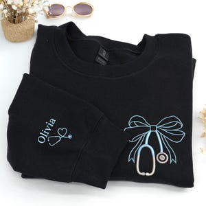 Könnte beinhalten: Schwarzes Sweatshirt mit hellblau gesticktem Stethoskop-Schleifendesign. Der linke Ärmel hat den Namen "Olivia" und ein Stethoskop. Das Sweatshirt ist auf einer weißen Oberfläche gefaltet.
