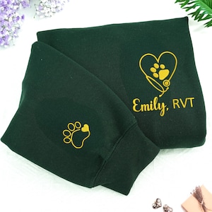 May include: Dark green sweatshirt with yellow embroidered paw prints and text. One paw print is on the sleeve. The other design features a heart with a paw print, stethoscope, and the name "Emily, RVT".