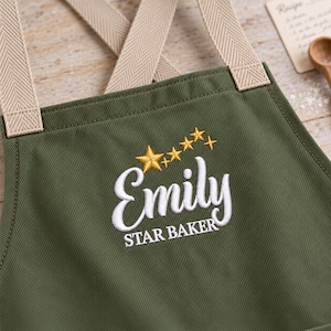May include: An olive green apron featuring beige herringbone straps and white embroidered text that reads Emily STAR BAKER, accented by a series of five gold embroidered stars of varying sizes positioned above the name.