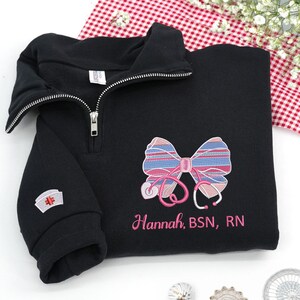 Könnte beinhalten: Schwarzes Sweatshirt mit Viertelreißverschluss und gesticktem Schleifen- und Stethoskop-Design. Die Schleife hat rosa und blaue Streifen. Der Text "Hannah, BSN, RN" ist rosa gestickt. Ein kleiner Krankenschwester-Kappen-Aufnäher befindet sich am Ärmel.