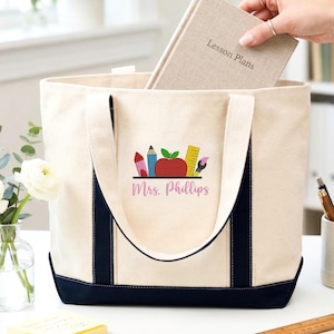 May include: Cream-colored canvas tote bag with navy blue trim and handles. The bag features a colorful graphic of school supplies and the name "Mrs. Phillips" in pink. A beige notebook labeled "Lesson Plans" is being placed inside the bag.