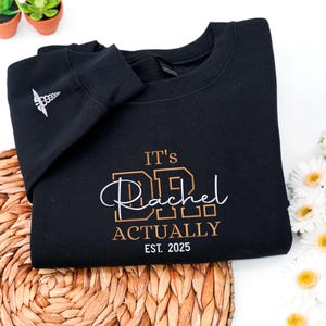 May include: Black sweatshirt with the text "IT'S Dr. Rachel ACTUALLY EST. 2025" in gold and white lettering. A medical symbol is on the left sleeve. The sweatshirt is on a woven mat with flowers and plants in the background.