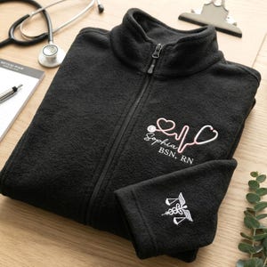May include: Black fleece zip-up jacket with a high collar. Features a pink and white embroidered stethoscope design with the name "Sophia" and credentials. A matching cuff displays a white caduceus symbol. A stethoscope and notepad are in the background.