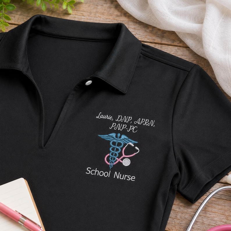 K&ouml;nnte beinhalten: Ein schwarzes Poloshirt mit wei&szlig;er Stickerei, die Laurie, DNP, APRN, PNP-PC und School Nurse liest. Unter dem Namen ist ein blaues Caduceus-Symbol mit einem rosa Stethoskop verschlungen, alles auf den linken Brustbereich des Shirts gestickt.
