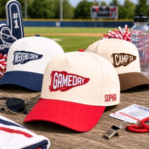 May include: Three baseball caps with embroidered pennant designs. One is white with a navy brim and 'WEEKEND CREW' text. One is cream with a red brim, 'GAMEDAY' text, and 'SOPHIA' on the side. One is cream with a brown brim and 'CAMP 24' text.