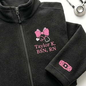 May include: Black fleece zip-up jacket with a pink bow and stethoscope design. The jacket has the name "Taylor K. BSN, RN" embroidered in pink. A pink bandage patch with a heart is on the sleeve. A stethoscope is in the background.