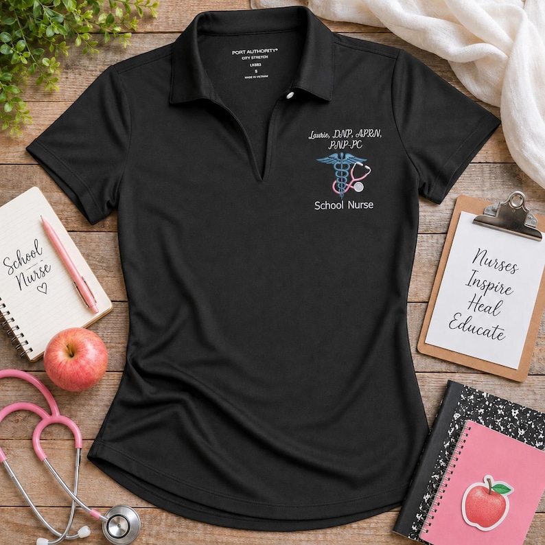 A black women’s polo shirt features a personalized school nurse design embroidered on the left chest.
The design includes the name and nursing credentials in white script above a blue medical symbol with a pink stethoscope.
“School Nurse” is stitched underneath in white thread for a clear professional finish.
White, blue, pink, and gray embroidery threads contrast sharply against the black fabric.
