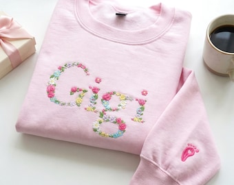 Könnte beinhalten: Hellrosa Sweatshirt mit dem Namen "Gigi" in bunten Blumen gestickt. Ein kleiner rosa Fußabdruck ist auf dem Ärmel gestickt. Eine Geschenkbox und eine Tasse Kaffee befinden sich im Hintergrund.