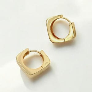 May include: Two gold-colored square hoop earrings. The earrings have a rounded square shape and a hinged closure. The earrings are displayed on a white surface. The earrings are a type of jewelry.