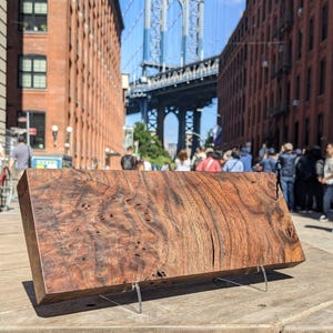 May include: A rectangular, polished wooden block with a rich, swirling grain pattern, displayed on clear acrylic stands. The wood's color ranges from deep brown to reddish hues. The background features a city street with a bridge.