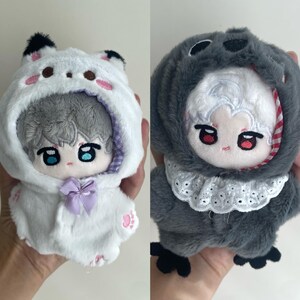 May include: Two plush dolls featuring embroidered facial features. The left doll has gray hair and a white hooded outfit with black ear tips, pink paw prints, and a lavender bow. The right doll has white hair and a dark gray hooded outfit with a white lace collar.