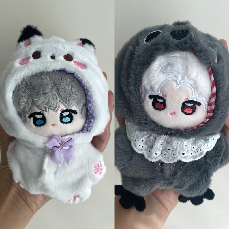 May include: Two plush dolls featuring embroidered facial features. The left doll has gray hair and a white hooded outfit with black ear tips, pink paw prints, and a lavender bow. The right doll has white hair and a dark gray hooded outfit with a white lace collar.