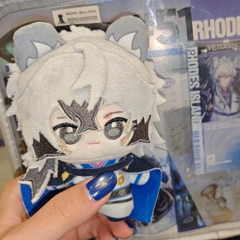 May include: A plush doll with a white fur-like head and gray ears. The doll has embroidered facial features and is wearing a blue and white outfit. A card with the text "RHODES ISLAND SILVERASH" is visible in the background.
