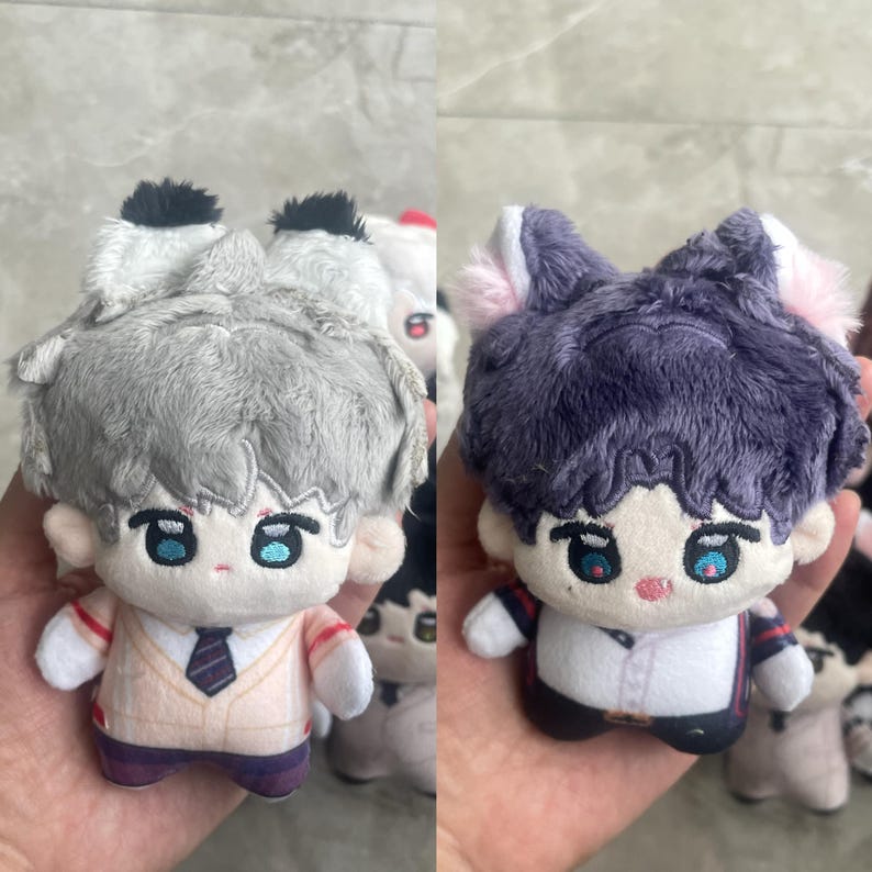 May include: Two plush dolls with embroidered facial features and animal ears. The left doll has gray hair, black-tipped ears, and a beige vest with a tie. The right doll has dark purple hair, pink-tipped ears, and a white shirt with dark accents.