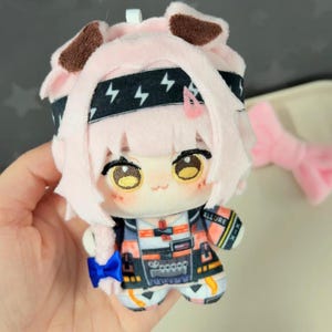 May include: A plush doll with pink hair and a black headband with lightning bolt designs. The doll has large, detailed eyes and is wearing a colorful outfit with the word "ALLURE" on the sleeve. A pink bow is visible in the background.