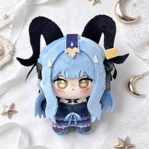 May include: A plush doll featuring a character with light blue hair and black horns. The doll has a detailed face with large eyes and is wearing a dark blue outfit. The doll is a collectible item.