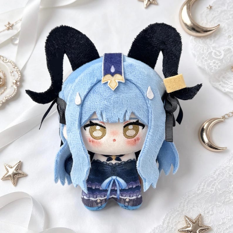 May include: A plush doll featuring a character with light blue hair and black horns. The doll has a detailed face with large eyes and is wearing a dark blue outfit. The doll is a collectible item.