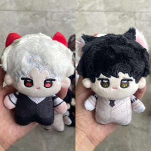 May include: Two plush dolls featuring embroidered facial features. The left doll has white hair, red horns, and a black vest over a striped shirt. The right doll has black hair, pink cat ears, and a light beige suit with a dark tie.