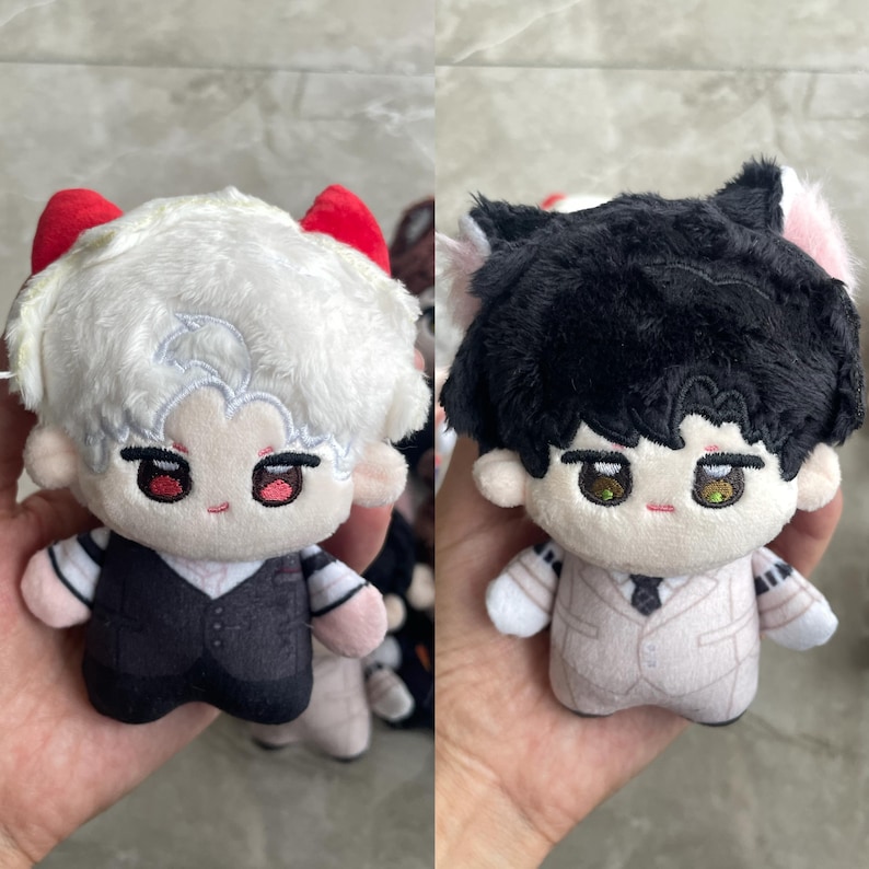 May include: Two plush dolls featuring embroidered facial features. The left doll has white hair, red horns, and a black vest over a striped shirt. The right doll has black hair, pink cat ears, and a light beige suit with a dark tie.
