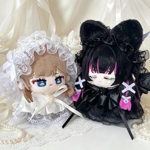 May include: Two plush dolls side by side. The left doll has light brown hair, blue eyes, and wears a white ruffled bonnet and dress with a pearl accent. The right doll has black hair with pink streaks, cat ears, and a black ruffled dress.