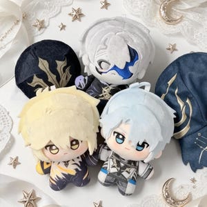 Puede incluir: Tres muñecos de peluche con cabello estilizado y rasgos faciales bordados. Un muñeco tiene cabello blanco y una máscara azul, otro tiene cabello rubio con detalles amarillos y otro tiene cabello azul claro. Visten trajes detallados con patrones dorados y azules.