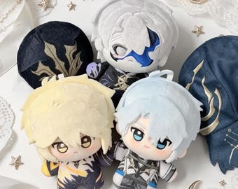 May include: Three plush dolls with stylised hair and embroidered facial features. One doll has white hair and a blue mask, one has blonde hair with yellow accents, and one has light blue hair. They wear detailed outfits with gold and blue patterns.