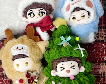 May include: Four plush dolls dressed in holiday costumes. One wears a red Santa suit, one a blue snowman hood, one a tan reindeer outfit with antlers, and one a green Christmas tree costume decorated with small ribbons and pom-poms.
