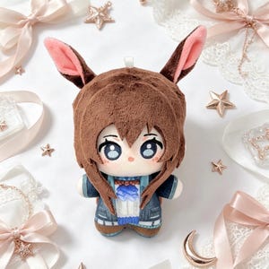 May include: A plush doll with brown hair and bunny ears, wearing a blue and white outfit. The doll has large, expressive eyes and is surrounded by decorative ribbons and star-shaped ornaments.