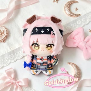 May include: A plush doll with pink hair, brown ears, and a black headband with lightning bolt designs. The doll wears a colorful outfit with a blue bow. The image includes decorative bows, a crescent moon, and the text "Goldenglow".