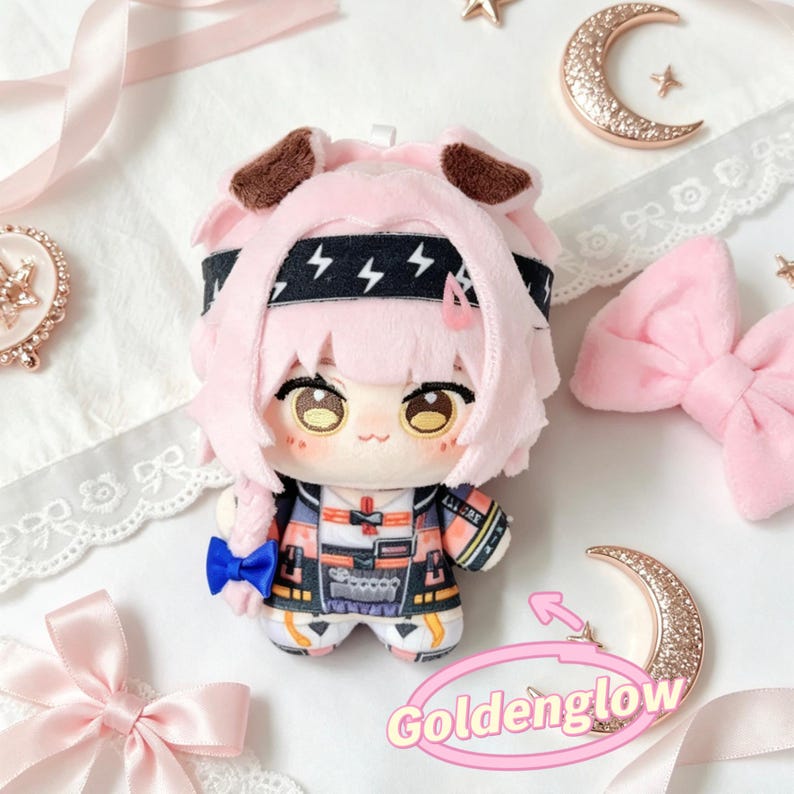 May include: A plush doll with pink hair, brown ears, and a black headband with lightning bolt designs. The doll wears a colorful outfit with a blue bow. The image includes decorative bows, a crescent moon, and the text "Goldenglow".