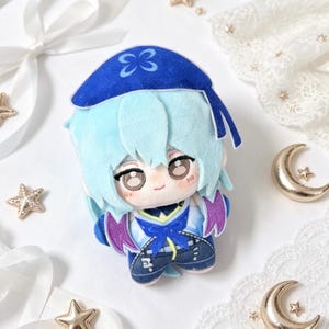 May include: A plush doll featuring a character with light blue hair and a blue hat with a butterfly design. The doll has a detailed outfit with blue, purple, and white accents. The doll is on a white surface with decorative stars and crescent moons.