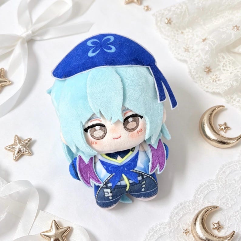 May include: A plush doll featuring a character with light blue hair and a blue hat with a butterfly design. The doll has a detailed outfit with blue, purple, and white accents. The doll is on a white surface with decorative stars and crescent moons.