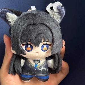 May include: A plush doll with a dark gray head and hair, blue eyes, and a detailed outfit. The doll has a small, white tag attached to the top. The doll is held in a person's hand.