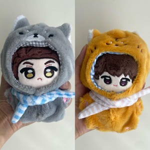 May include: Two plush dolls with embroidered facial features and brown hair. One doll wears a gray animal-themed hood with a blue and white striped scarf. The other doll wears an orange animal-themed hood with a pink and white striped scarf.