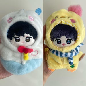 May include: Two plush dolls with embroidered facial features and dark hair. The left doll wears a white and light blue hooded outfit with a red and yellow accent. The right doll wears a yellow hooded outfit with a pink pom-pom and blue scarf.