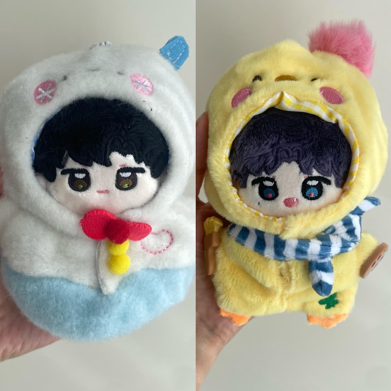 May include: Two plush dolls with embroidered facial features and dark hair. The left doll wears a white and light blue hooded outfit with a red and yellow accent. The right doll wears a yellow hooded outfit with a pink pom-pom and blue scarf.