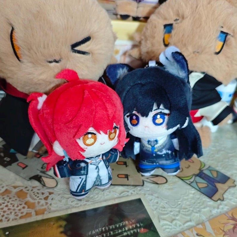 May include: Two plush dolls with anime-style features. One doll has red hair and the other has black hair. Both dolls are wearing detailed outfits. Two larger plush toys are in the background.