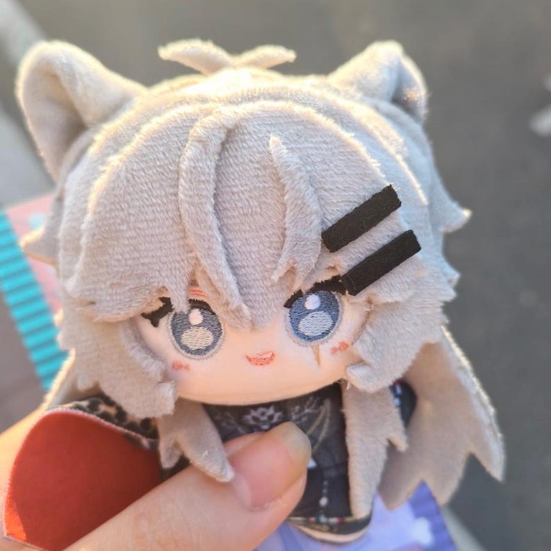 May include: A plush doll with light gray hair and cat ears. The doll has blue eyes, a small red mouth, and two black barrettes. The doll is wearing a dark outfit with a floral pattern.