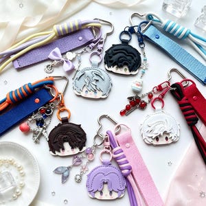 May include: A collection of six anime-style character head keychains, each attached to a fabric wrist strap. The keychains feature diverse hair colors including black, silver, brown, and purple, with various charms like bows, beads, and metal shapes.
