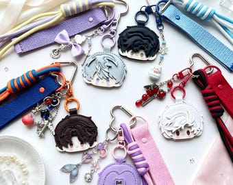 May include: A collection of six anime-style character head keychains, each attached to a fabric wrist strap. The keychains feature diverse hair colours including black, silver, brown, and purple, with various charms like bows, beads, and metal shapes.