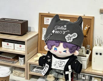 May include: A plush doll with purple hair and a black hat with the text "Follow meow." The doll is dressed in a black jacket, a white t-shirt with text, and denim trousers. The doll is on a desk with a speaker, a corkboard, and other items.