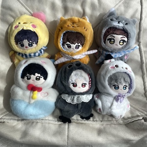 May include: Six plush dolls dressed in various animal-themed hooded outfits. The dolls feature embroidered facial details, including large eyes and hair. The outfits include a yellow chick, orange fox, gray wolf, white seal, dark gray bird, and white cat.
